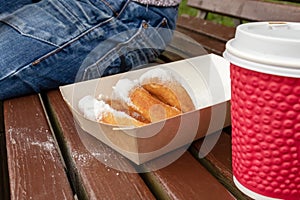 Closeup of beignet pastries, donuts in a paper takeaway box and a cup of coffee on a bench in park. The delicious
