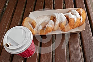 Closeup of beignet pastries, donuts in a paper takeaway box and a cup of coffee on a bench in park. The delicious deserts covered