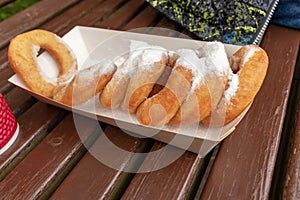 Closeup of beignet pastries, donuts in a paper takeaway box and a cup of coffee on a bench in park. The delicious deserts covered