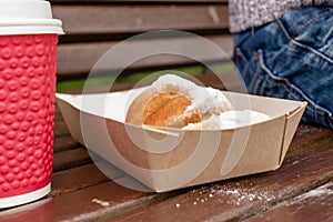 Closeup of beignet pastries, donuts in a paper takeaway box and a cup of coffee on a bench in park. The delicious deserts covered