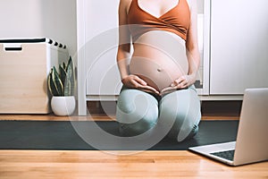 Closeup belly of pregnant woman practicing yoga online with laptop