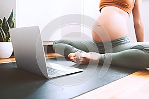 Closeup belly of pregnant woman practicing yoga online with laptop