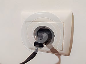 Closeup of a black electrical plug inserted into a creamcolored wall socket. Simple, clean image perfect for illustrating photo