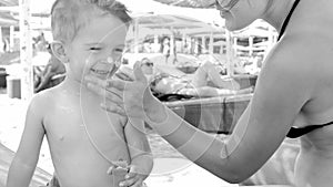 Closeup black and white cheerful portrait of little toddler boy smiling while mother applying sunscreen lotion on his