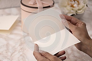 Closeup of blank greeting card.Woman holding postcard against present box with pink bow,white flower on the desk.Free space