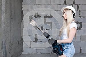 Closeup blonde girl foreman in white construction helmet holding professional perforator, drill in house under construction.