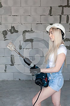 Closeup blonde girl foreman in white construction helmet holding professional perforator, drill in house under construction.