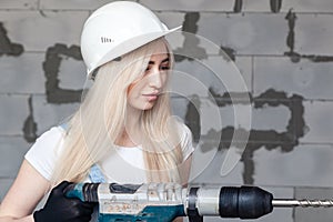 Closeup blonde girl foreman in white construction helmet holding professional perforator, drill in house under construction.