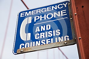 Closeup of a blue [Emergency Phone And Crisis Counseling] sign on a bridge with a blurry background