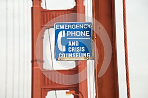 Closeup of a blue [Emergency Phone And Crisis Counseling] sign on a bridge with a blurry background