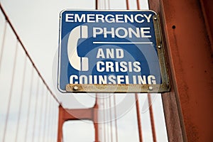 Closeup of a blue [Emergency Phone And Crisis Counseling] sign on a bridge with a blurry background