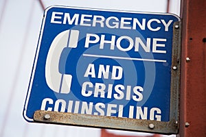 Closeup of a blue [Emergency Phone And Crisis Counseling] sign on a bridge with a blurry background
