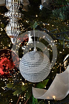 Silver white ornament hanging from greenery and surrounded by additional red and silver ornaments and ribbon.