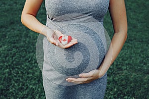 Closeup body of white Caucasian pregnant woman touching her belly and holding wooden heart with Canadian flag