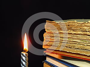 Closeup of books pile and a burning candle. A pile of old books is pictured against dark black background.