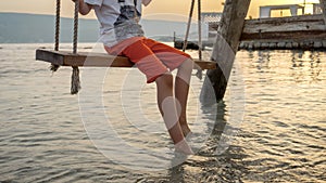 Closeup of boy& x27;s feet touching sea waves while swinging on swing at the ocean beach on sunset. Holiday, summer