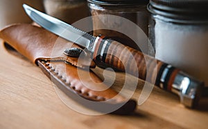 Closeup of a brown knife with a case surrounded by spice jars on a wooden table under the lights