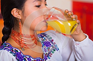 Closeup brunette woman with red necklace and earrings, drinking orange juice from glass, closing her eyes enjoying