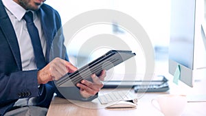 Closeup of businessman holding tablet, sitting and working at desktop computer in modern startup. Professional in office