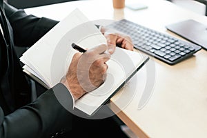A closeup of the businessman's hands jotting something in a small notebook while looking at a computer monitor at his desk
