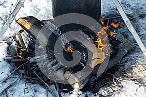 Closeup campfire in the snow and caked pot in soot over the fire on tripod, winter outdoor cooking at the campsite