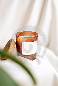 Closeup of candle in glass jar with blank label. Blurred green plant, white silk backgound. Front view. Handmade