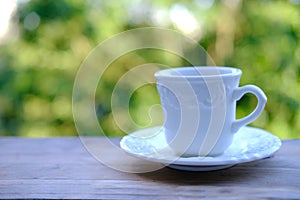 Closeup of cappuccino coffee in white cup and saucer on wooden table, beautiful blurred natural background with green foliage,