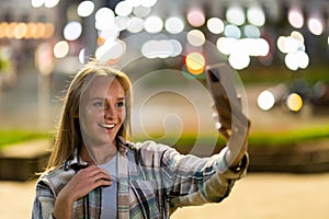 Closeup cheerful girl taking selfie photo by mobile phone on night city street. Portrait of happy woman showing victory sign to
