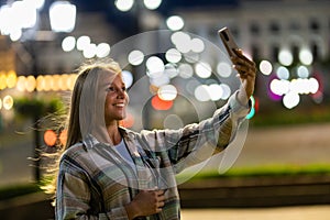 Closeup cheerful girl taking selfie photo by mobile phone on night city street. Portrait of happy woman showing victory sign to