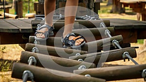 Closeup of childs' feet walking over wobbly rope bridge at adventure park in summer camp. Kids sports, summer holiday, fun