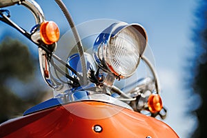 Closeup chrome detail and headlamp of orange retro vintage scooter under blue sky and sun on the blurred background.