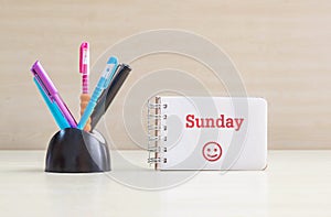 Closeup color pen with black ceramic desk tidy for pen and red sunday word in white page and happy face emotion at the note book o