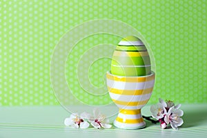 Closeup colorful painted Easter egg in vibrant modern egg stand surrounded by white flowering tree branches on green background