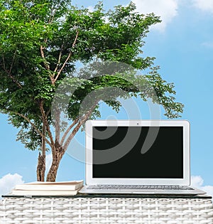 Closeup computer on blurred wood weave table and book on tree and blue sky with cloud background , beautiful work place at the