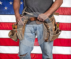 Closeup carpenter holding hammer and wearing worn out old, leather tool belt with hand tools in front of American flag photo