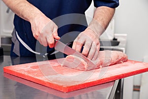 Closeup cook hand cuts pork fillet steak with sharp knife on red plastic cutting board on metal table in restaurant kitchen.