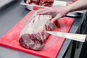 Closeup cook`s hand cuts ribeye marbled beef steak with sharp knife on red plastic cutting board on metal table in restaurant