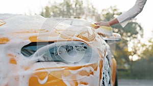 Closeup cropped image of hand of young caucasian woman with yellow sponge washing hood and headlights of her car at a