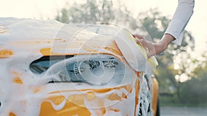 Closeup cropped image of hand of young caucasian woman with yellow sponge washing hood and headlights of her car at a