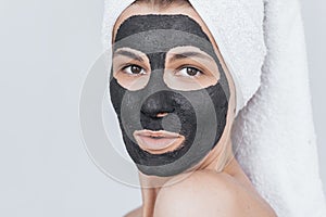 Closeup cropped studio portrait of amazed young woman with black clay organic mask on her face, wears white towel on hair. Female