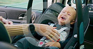 Closeup crying baby boy sitting in the baby car seat inside of car Mother fastens safety belts lifts the handle and