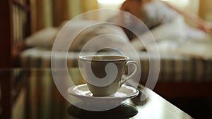 Closeup cup of hot tea or coffee, young woman waking up stretching arms on the bed in the morning. blurred focus