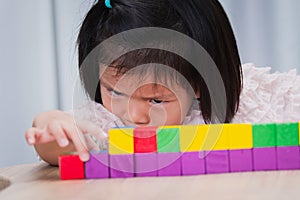 Closeup cute Asian girl playing with colorful wooden cube. Child intently aims at placement of wooden blocks.