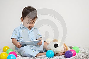 Closeup cute asian kid look at the tablet at home on gray carpet with doll and colorful ball and cement wall textured background w