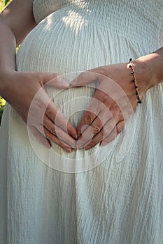 Closeup cute composition with a pregnant young woman wearing white dress makes a heart shaped symbol with her hands on her belly.