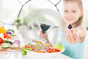 Closeup cute little girl making salad. Holding olive. Child cooking.