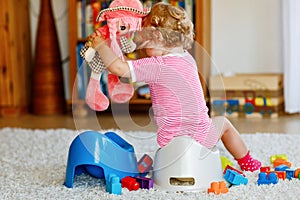 Closeup of cute little 12 months old toddler baby girl child sitting on potty. Kid playing with doll toy. Toilet