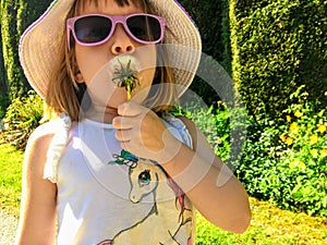 A closeup of cute 5 year old girl holding and blowing on a dandelion in a garden.  She is wearing a unicorn shirt