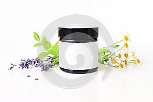 Closeup of dark glass jar with blank paper label. Mint leaves, lavender and feverfew flowers on white table backround photo