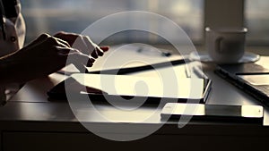 Closeup, dark silhouette of female hands. she is typing something in tablet, next is a cup, mobile, laptop on table